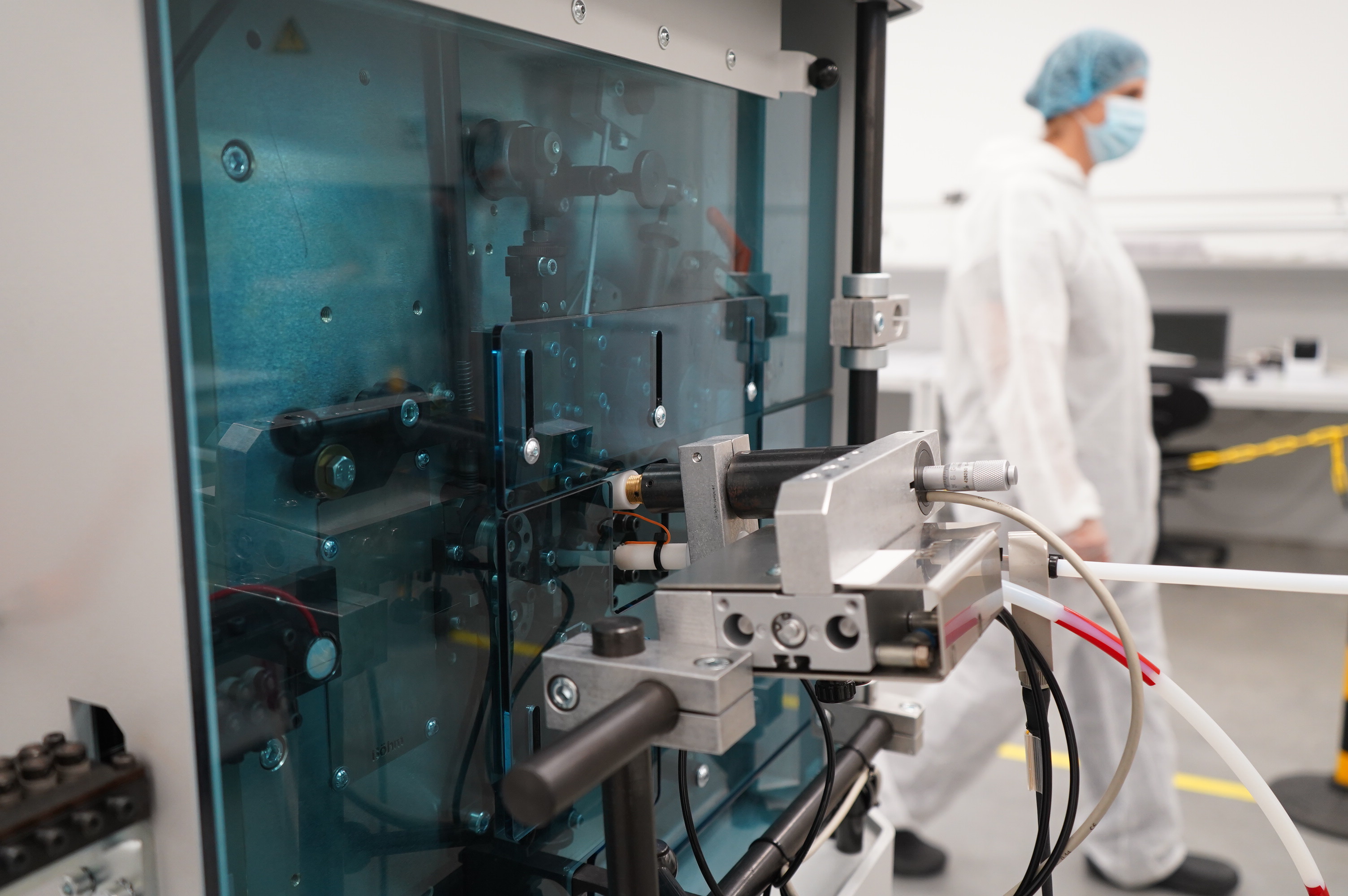 Automated manufacturing machine with transparent safety panels in a cleanroom environment, with a technician wearing protective clothing in the background.