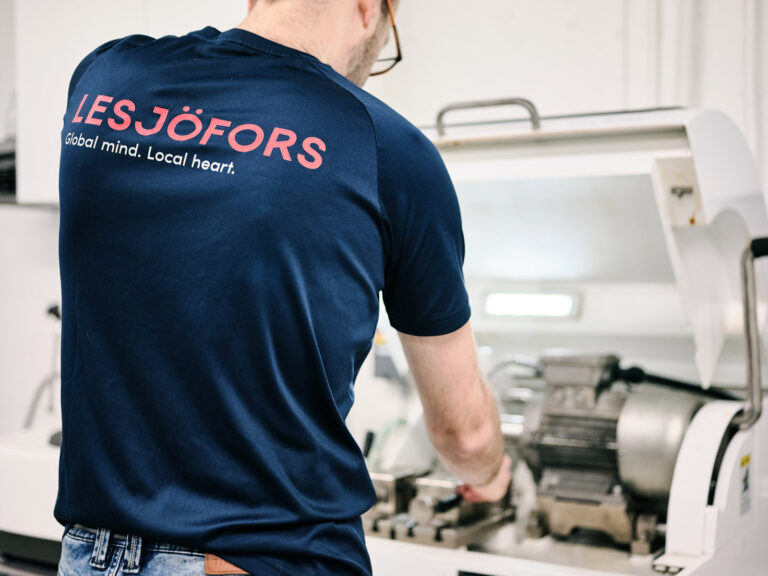 A male Lesjofors team member wearing a dark blue branded T-shirt with red writing that says 'Lesjofors - Global mind. Local Heart' manufacturing springs.