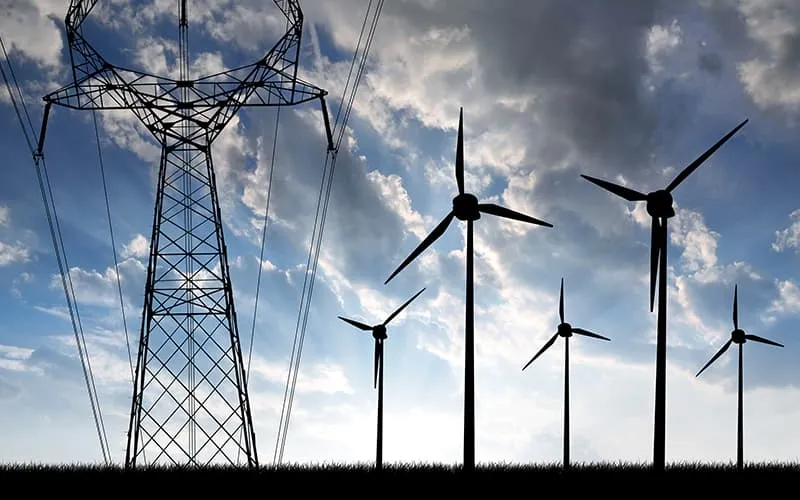 Silhouettes of wind turbines and an electricity pylon against a blue sky, showcasing the role of Lesjöfors’ springs within wind energy generation.